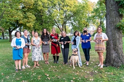 [ai] A group of nine women standing in a park, each holding an award. The scene includes a yellow dog sitting in front of them, with trees and green grass in the background.