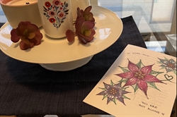 [ai] A decorative display featuring a white ceramic cup with floral designs, alongside small artificial flowers on a round white plate. A colorful card with hand-drawn flowers reads, 'YOU ARE NOT ALONE.'
