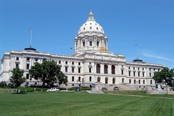 [ai] The Minnesota State Capitol building, featuring a grand dome and ornate architectural details, set against a clear blue sky. The front lawn is green with trees and vehicles visible nearby.