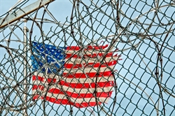 American flag pictured behind barbwire and a chain-link fence American flag pictured behind barbwire and a chain-link fence