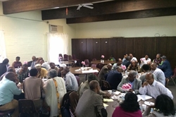 [ai] A large group of people seated at round tables in a well-lit room, engaged in discussion. Some individuals are taking notes while others are conversing. Centerpieces with flowers adorn the tables.