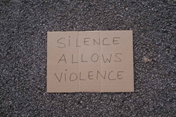 [ai] A cardboard sign with handwritten text reading "SILENCE ALLOWS VIOLENCE" placed on a textured asphalt surface.