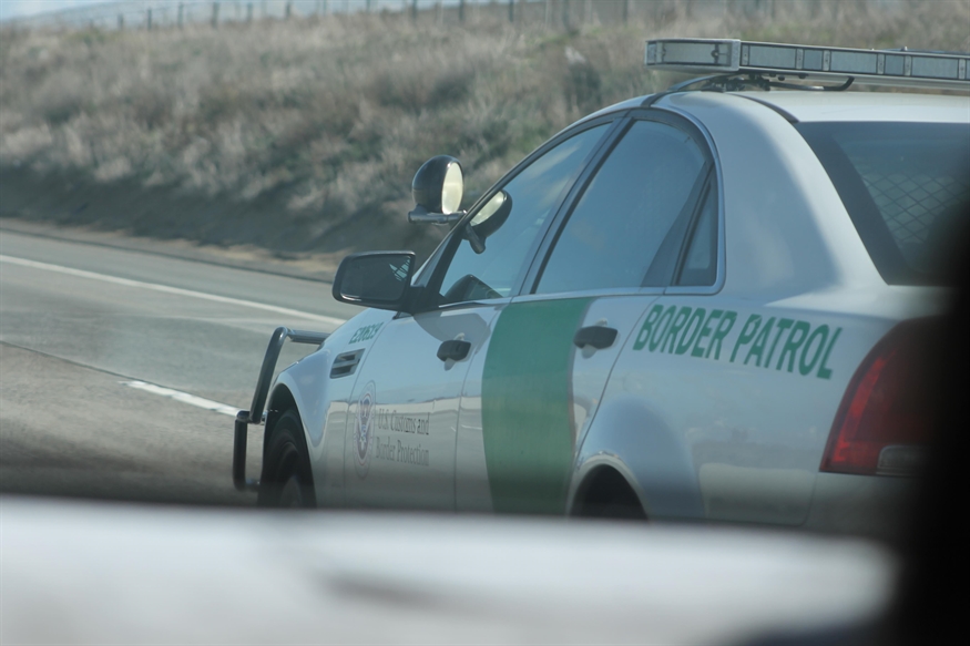 [ai] A U.S. Border Patrol vehicle parked on the side of a highway. The car features a green stripe and the words 'BORDER PATROL' on the side, with a blurred view of another vehicle in the foreground.