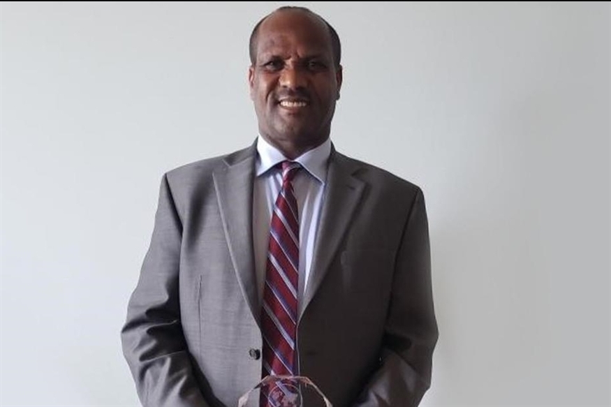 [ai] A man in a suit and tie standing against a plain background, smiling while holding a crystal award in his hands.