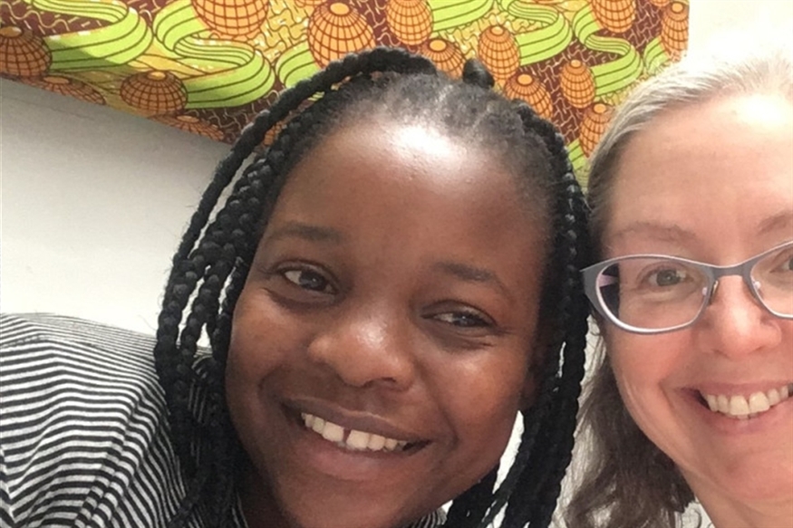 [ai] Two women posing for a selfie, smiling at the camera. One woman has braided hair and is wearing a striped shirt, while the other has glasses and light-colored hair. A colorful patterned background is visible behind them.