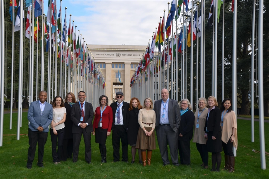 [ai] A diverse group of professionals standing together outside the United Nations building in Geneva, surrounded by flags from various countries. The scene captures a formal gathering with a mix of attire.