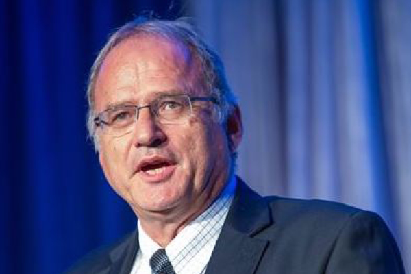 [ai] A man speaking at a podium, wearing glasses, a black suit, and a light blue shirt with a patterned tie. The backdrop features soft blue and white drapes.