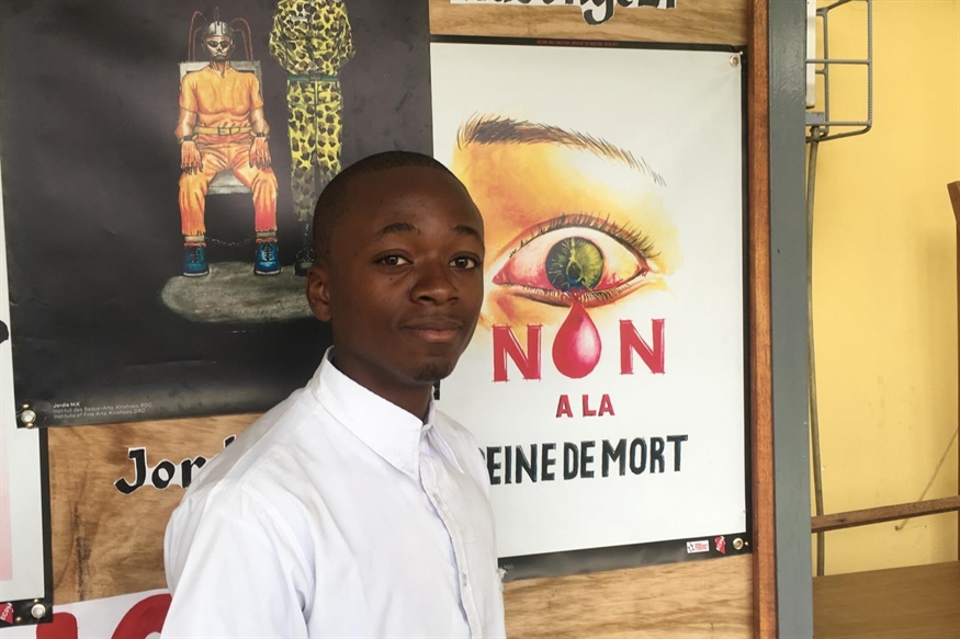 [ai] A young man in a white shirt standing against a poster displaying an eye with a teardrop and the text "NON A LA REINE DE MORT." Behind him, there is also a colorful poster of a figure in a creative costume.