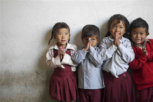 [ai] Four children standing in a line against a white wall, with their hands together in a prayer position. They are wearing school uniforms, consisting of shirts and skirts or shorts in various colors.