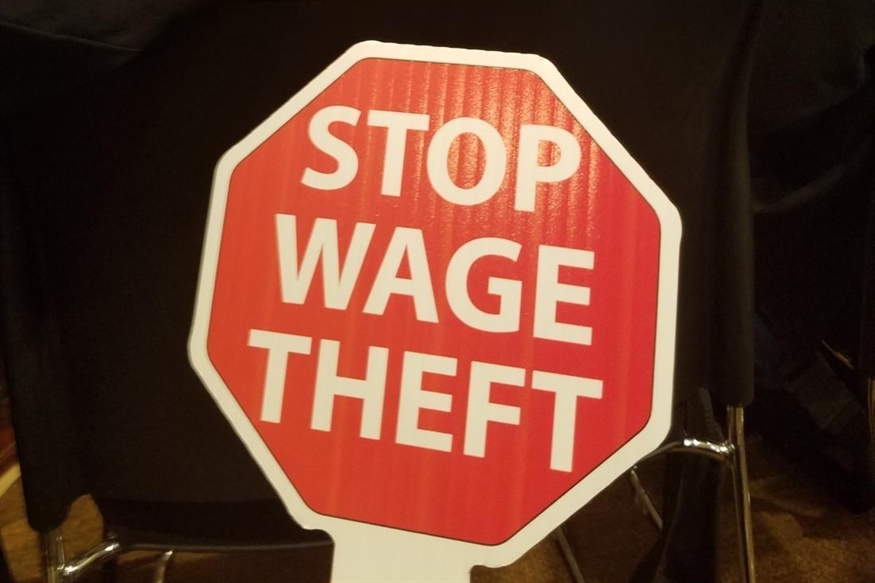 [ai] A stop sign with the message "STOP WAGE THEFT" written in bold white letters on a red background, placed on a table in a public setting.
