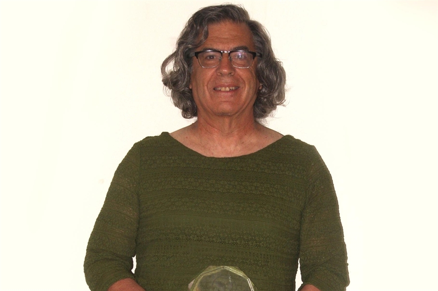 [ai] A person with shoulder-length curly hair, wearing a green top, holding a clear award in front of a plain white background. The individual is smiling and appears to be in a celebratory context.