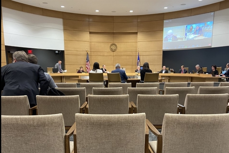 [ai] A view of a legislative meeting room with empty chairs in the foreground and several individuals seated around a circular table. A screen displays presentations, and flags are visible in the background.